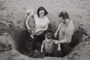 A black and white photo shows a mother, father, and two young children playing in a sand pit. The light is soft and creates a moody atmosphere.