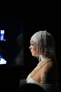 Photo of a model in a white dress and pearl-covered headwear, taking a breath backstage at the Vancouver Indigenous Fashion Week, just before going on the runway.