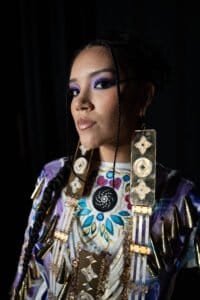 Photo of a model backstage at the Vancouver Indigenous Fashion Week, illuminated by a phone light, showcasing beautiful jewelry.