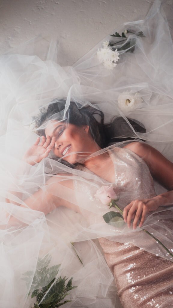 High-fashion editorial photo of model Kwan Scherp lying on her back amidst pink roses and tulle, with a joyful smile, at Shoreline Studios Vancouver, photographed by Yar Tareen (Zur Photography).