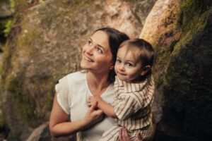 A mother and toddler stand in a sun-dappled forest clearing. The mother looks down at her child, both smiling as they gaze up at the tall trees.